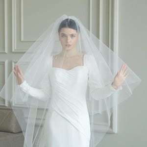 Puede incluir: Una novia con un vestido de novia blanco con mangas largas y una cola fluida. Lleva un velo transparente que le cubre la cabeza y los hombros. El vestido tiene un escote cuadrado y un corpiño ajustado.