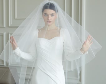 May include: A bride in a white wedding dress with long sleeves and a flowing train. She is wearing a sheer veil that drapes over her head and shoulders. The dress has a square neckline and a fitted bodice.