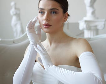 May include: A woman in a white strapless dress and long white gloves. The gloves reach past the elbows. The background features white statues and a white sofa.