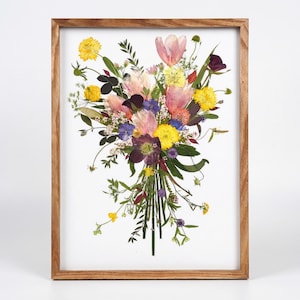 May include: A framed wall art piece featuring a pressed flower arrangement. The bouquet includes yellow, pink, and deep purple blossoms with green stems and foliage, all displayed against a white background within a light brown wooden frame.