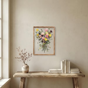 May include: A rectangular wooden frame containing a pressed flower arrangement featuring yellow, pink, purple, and white blossoms with green stems and leaves. The floral display is arranged in a vertical bouquet shape against a light background.