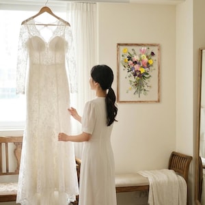 May include: A floor-length white lace wedding dress with long sleeves, a sweetheart neckline, and a sheer lace bodice. The dress features intricate floral embroidery throughout the fabric, a fitted waist, and a flowing skirt that reaches the floor.
