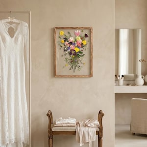 May include: A white lace long-sleeve dress hangs on a rack next to a rectangular wooden frame containing a pressed flower bouquet with yellow, pink, and purple blossoms. A wooden bench with folded light-colored fabric sits below the wall art.