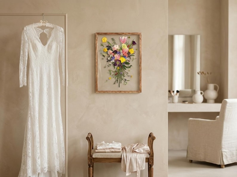 May include: A white lace long-sleeve dress hangs on a rack next to a rectangular wooden frame containing a pressed flower bouquet with yellow, pink, and purple blossoms. A wooden bench with folded light-colored fabric sits below the wall art.