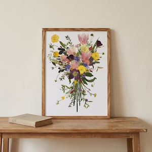 May include: A rectangular wooden frame containing a print of a pressed flower bouquet. The arrangement features yellow, pink, purple, and white blossoms with green stems and leaves, all set against a plain white background within the frame.