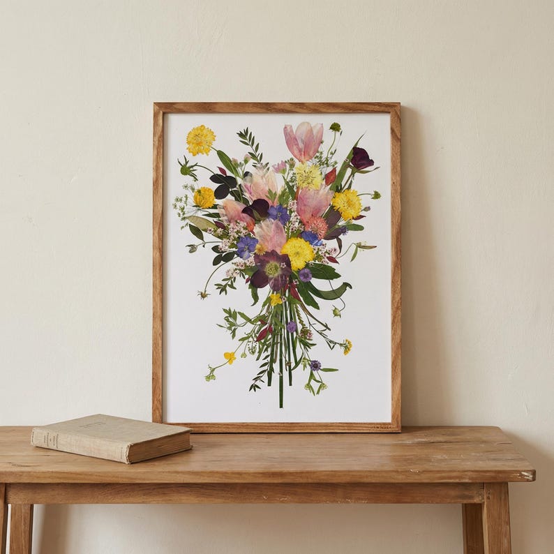 May include: A rectangular wooden frame containing a print of a pressed flower bouquet. The arrangement features yellow, pink, purple, and white blossoms with green stems and leaves, all set against a plain white background within the frame.