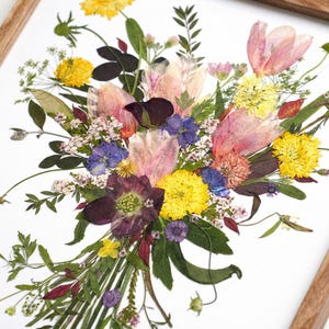 May include: A framed wall art piece featuring a pressed flower arrangement on a white background. The bouquet includes yellow, pink, purple, and white dried blossoms with green foliage, all encased in a light-toned wooden frame.