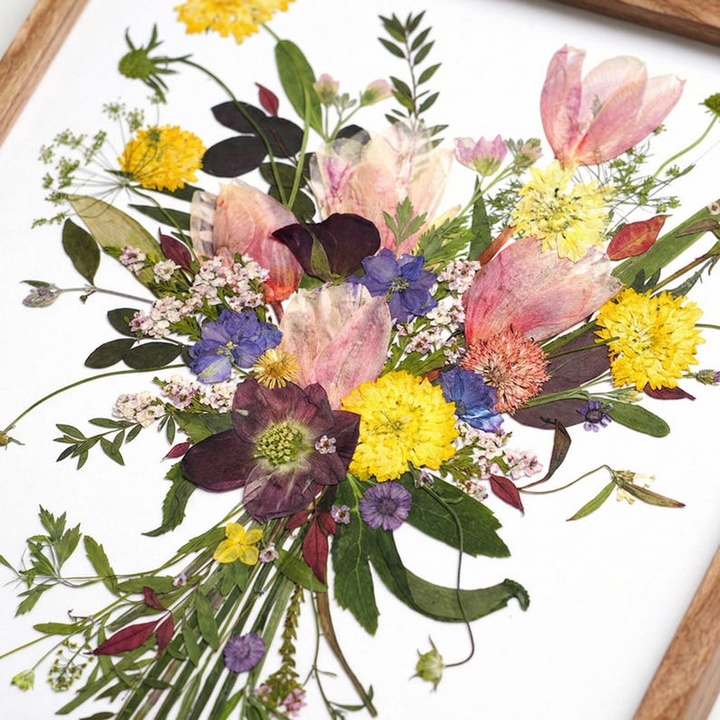 May include: A framed wall art piece featuring a pressed flower arrangement on a white background. The bouquet includes yellow, pink, purple, and white dried blossoms with green foliage, all encased in a light-toned wooden frame.