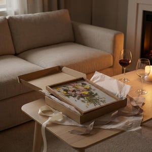 May include: A rectangular wooden frame containing a pressed flower arrangement with yellow, pink, and purple petals and green foliage, resting inside an open cardboard gift box with white tissue paper and a light-colored ribbon on a wooden table.