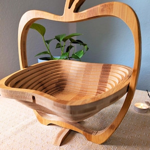 Op de afbeelding: Een houten fruitmand in de vorm van een appel met een inklapbaar ontwerp. De mand is gemaakt van natuurlijk bamboe en heeft een lichtbruine kleur.