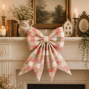 K&ouml;nnte beinhalten: Eine gro&szlig;e, dekorative Stoffschleife mit einem Patchwork-Design aus zartrosa, cremefarbenen und gedeckten gr&uuml;nen Karos mit feinen Blumenmustern. Die Schleife besteht aus einem zentralen, geknoteten Stoffband und zwei langen, spitz zulaufenden Enden.