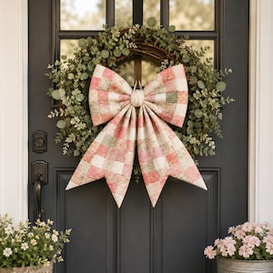 May include: A round wreath made of intertwined brown twigs and green eucalyptus leaves, featuring a large, central fabric bow with a pink, white, and sage green checkered pattern. The bow has two wide top loops and two long, pointed tails hanging downward.