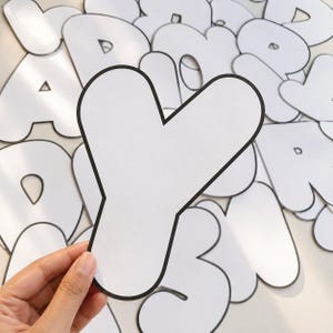 May include: A large, white, die-cut letter Y with a thick black outline. The letter is presented against a background of various other white, black-outlined letters and numbers, creating a collection of individual paper cutouts for educational use.