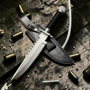 May include: A survival knife with a silver blade and black handle rests in a black leather sheath. The knife is on a weathered wooden surface, surrounded by brass bullet casings and small gold-colored fragments.