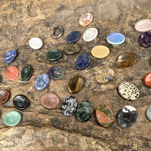 May include: A collection of 25 oval-shaped polished stones in various colors and patterns, including green, red, blue, brown, black, white, and gray. The stones are arranged on a wooden surface.
