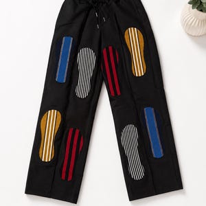 May include: Black trousers featuring an elastic waistband with a drawstring and multiple oval-shaped patches. The patches display vertical stripes in blue, white, red, and mustard yellow, arranged in a scattered pattern across the front legs.