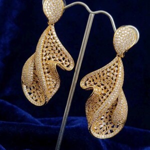 May include: A pair of gold earrings with a leaf-shaped design. The earrings are embellished with small, sparkling stones and have an open, intricate pattern. The earrings are displayed against a dark blue background.