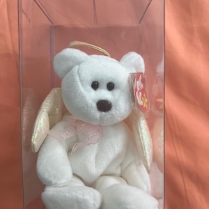 Könnte beinhalten: Weißer Plüsch-Engelbär mit goldenem Heiligenschein und schillernden Flügeln. Der Bär sitzt in einer durchsichtigen Plastikvitrine.