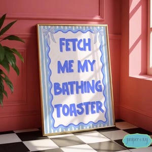 Könnte beinhalten: Ein gerahmter Druck mit dem Text "FETCH ME MY BATHING TOASTER" in Blau auf weißem Hintergrund, goldfarben gerahmt. Der Druck hat einen hellblauen Rand mit Schleifenakzenten. Der Druck lehnt an einer rosa Wand auf einem schwarz-weiß karierten Boden.