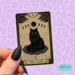 May include: A rectangular tarot card featuring a black cat sitting on a dark cloud. Above the cat are moon phases and stars. The card has a beige background with a black border and the text THE VOID at the bottom, with the Roman numeral II at the top.