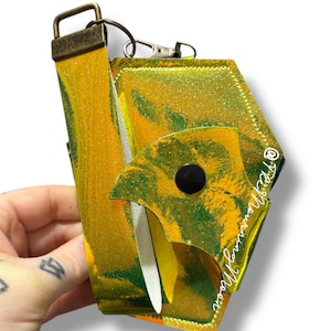 May include: An iridescent wallet with a yellow and green marbled pattern. The wallet features a black button closure and a wrist strap with a bronze-coloured metal clasp. The text "@TheMorningMoon" is visible on the wallet.