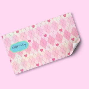 May include: A pink and white argyle pattern with small pink hearts scattered throughout. The design includes a light blue label with the text "juniper & ivy". The corner of the paper is folded over.