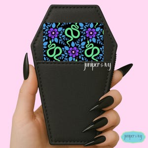 May include: Black coffin-shaped cardholder with a floral and snake design. The cardholder has a black exterior and a patterned interior with green snakes, purple flowers, and blue leaves on a black background. The brand name "juniper & ivy" is printed on the cardholder.