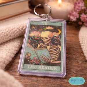 May include: A clear acrylic keychain featuring a tarot card design. The card depicts a skeleton reading a book, with the text "THE READER" at the bottom. The background is a starry night sky with a crescent moon.