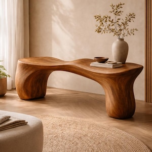 Puede incluir: Un banco de madera maciza con un diseño único y fluido. El banco es de color marrón cálido y tiene una parte superior curvada. Un jarrón con vegetación, un cuenco pequeño y libros están colocados sobre el banco. Una alfombra redonda de yute y un otomán beige están en primer plano.