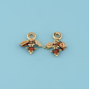 May include: Two gold-toned charms with a small figure wearing a hat and coat, with a camera and a small suitcase. The charms have a gold ring at the top for attaching to a bracelet or necklace. The background is a solid light blue.