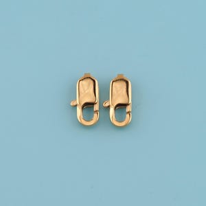 May include: Two gold-colored lobster claw clasps, each with a curved hook and a rectangular body. The clasps are displayed against a light blue background, showcasing their shiny, metallic finish. These clasps are likely used for jewelry making or crafting.