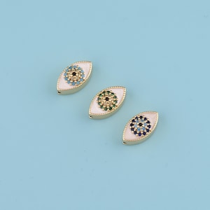 Könnte beinhalten: Drei goldfarbene, augenförmige Charms mit weißem Emaille und farbigen Edelsteinakzenten. Jeder Charm hat ein anderes Farbschema: blau, grün und blau-schwarz. Die Charms sind auf einer hellblauen Oberfläche angeordnet.