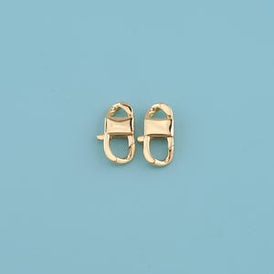 May include: Two gold-colored swivel clasps on a light blue background. Each clasp has a rectangular center piece and a curved hook at the top and bottom. The clasps are designed for attaching to bags or other accessories.