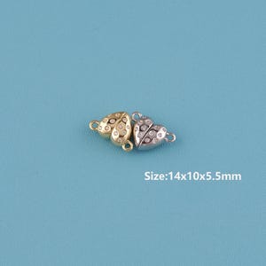 May include: Two heart-shaped jewelry clasps, one gold-toned and one silver-toned, each encrusted with small, clear crystals. The clasps are connected and have a size of 14x10x5.5mm. The background is a solid light blue.