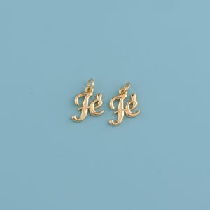 May include: Two gold-colored pendants spelling "Je Je" in a cursive font. Each pendant has a small loop at the top for attaching to a chain. The pendants are set against a light blue background.