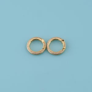 Puede incluir: Dos cierres redondos de color dorado con forma de anillo. Cada cierre está incrustado con pequeñas piedras preciosas transparentes. Los cierres están colocados sobre un fondo azul claro liso. Probablemente se utilizan para la fabricación de joyas o como elementos decorativos.