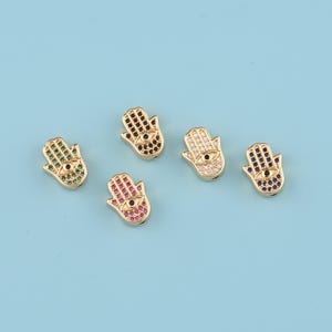 Puede incluir: Cinco amuletos dorados con forma de mano de Fátima, cada uno adornado con gemas de colores. Los amuletos presentan un diseño de ojo en el centro y están sobre un fondo azul claro. Las gemas son verdes, negras, blancas, rojas y azules.