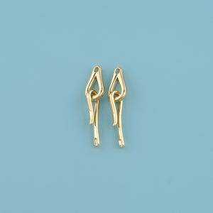 May include: Two gold-coloured, abstract-shaped earrings against a light blue background. Each earring has a looped top and a long, slender, dangling bottom. The earrings are symmetrical and appear to be made of metal.
