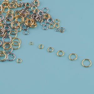 May include: A collection of gold and silver-colored jump rings of various sizes, scattered on a light blue surface. The rings are open, ready for use in jewelry making or other crafts. The image shows a variety of sizes and finishes.