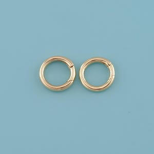 May include: Two gold-colored, circular spring clasp rings are displayed against a light blue background. The clasps are round with a hinged opening. The rings are likely used for jewelry making or crafting.