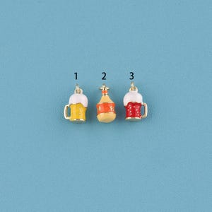 Könnte beinhalten: Drei Miniatur-Bier-Anhänger. Der erste und dritte Anhänger sind gelbe und rote Bierkrüge mit weißem Schaum. Der zweite Anhänger ist eine kleine orangefarbene Flasche. Alle drei Anhänger haben goldfarbene Metallakzente und sind vor blauem Hintergrund.