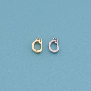 May include: Two metal jewelry clasps, one gold-toned and one silver-toned, are displayed against a solid blue background. The clasps are circular with a small loop at the top for attachment. They are designed for jewelry making.