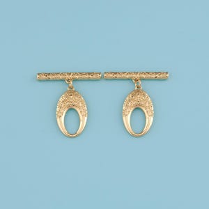 May include: Two gold-colored metal brooches. Each brooch has a horizontal bar with an ornate design, from which hangs an oval pendant with a central opening. The pendants are also decorated with intricate patterns. The background is a solid light blue.