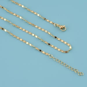 Op de afbeelding: Een delicate goudkleurige ketting met een herhalend ovaal schakelontwerp. De ketting heeft een karabijnsluiting en een verstelbare ketting. De sieraden worden gepresenteerd tegen een lichtblauwe achtergrond.