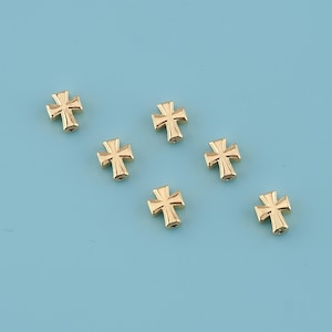 Puede incluir: Siete pequeños dijes de cruz dorados están dispersos sobre una superficie azul claro. Cada cruz tiene un diseño clásico con cuatro brazos de igual longitud. Las cruces parecen ser de metal y tienen un acabado brillante.