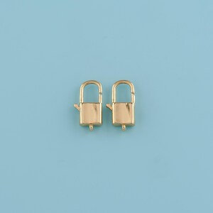 May include: Two gold-colored, rectangular-shaped clasp locks with arched handles. The locks are positioned side-by-side against a light blue background. Each lock has a small, protruding lever on one side and a small hole at the bottom.
