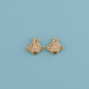 May include: Two gold-colored, fish-shaped charms with small wings and crowns. The charms are encrusted with small, clear crystals and have black eyes. The background is a solid light blue.