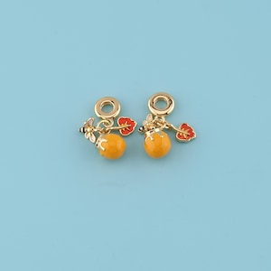 Puede incluir: Dos colgantes dorados con diseño de tarro de miel. Cada colgante presenta un tarro de miel redondo y naranja, una flor roja y una pequeña abeja. Los colgantes están suspendidos de un anillo dorado, perfectos para la creación de joyas.