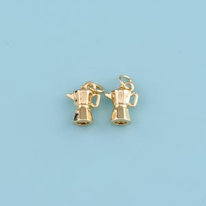 Puede incluir: Dos pequeños colgantes dorados con forma de cafetera, con un lazo en la parte superior para sujetarlos a un collar o pulsera. Los colgantes están detallados con un pico, un asa y una base, sobre un fondo azul claro.