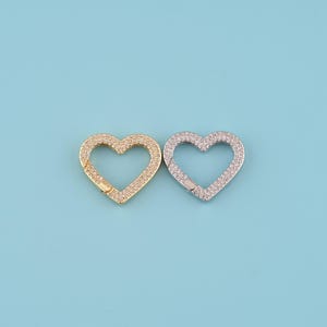 May include: Two heart-shaped clasps, one gold-toned and one silver-toned, each encrusted with small, sparkling stones. The clasps are open, revealing their locking mechanisms. The background is a solid light blue.